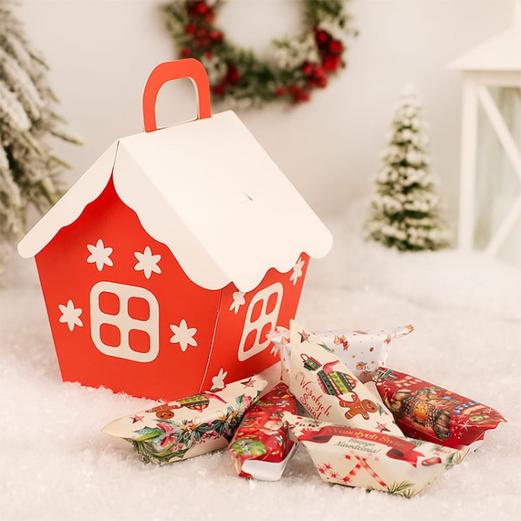 Czerwone pudełko w kształcie domku z białym dachem wypełnione świątecznymi krówkami z życzeniami.