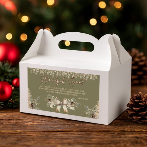 Białe pudełko świąteczne z rączką ozdobione napisem „Wesołych Świąt” na tle choinki.
