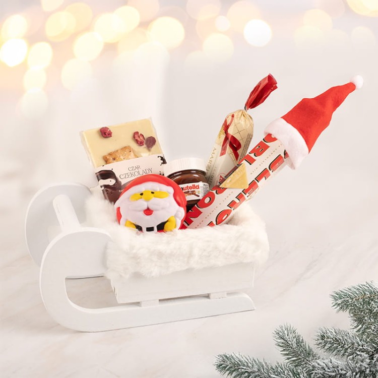 Zestaw świąteczny ze słodyczami dla dziecka w białych drewnianych saniach.